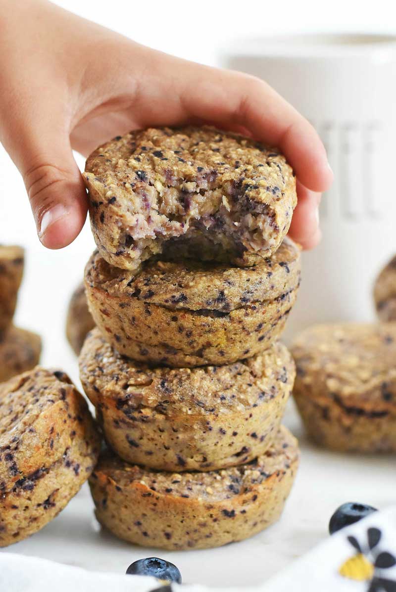 4 ingredient blueberry banana muffins are stacked 4 high. The top muffin is being held by a small white hand and has a bite taken out of it.