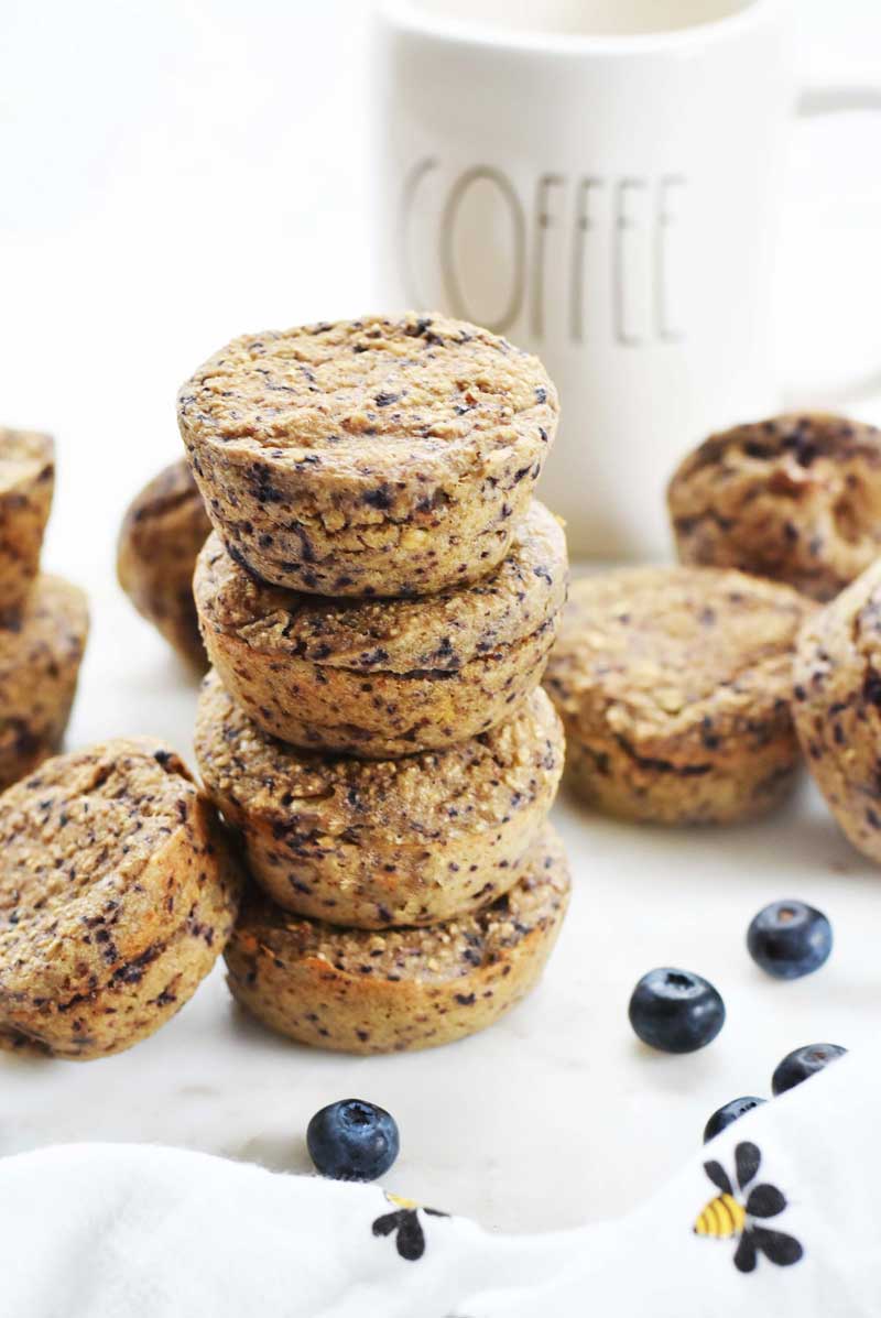 Blueberry Oatmeal Muffins are stacked 4 muffins high. There is a coffe mug to the right of the image, fresh blueberries, a bee napkin and some additional muffins scattered.