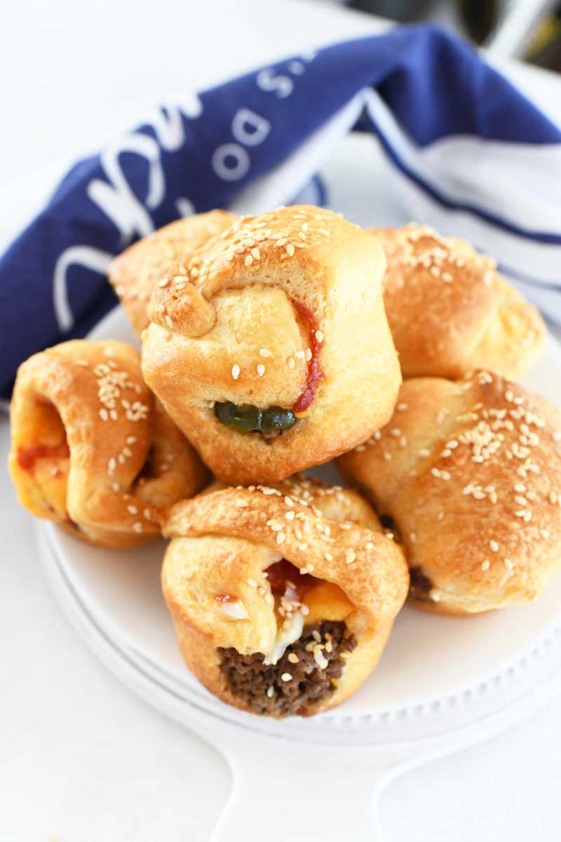 Cheeseburger Cups are made with crescent dough in a muffin tin. A blue and white napkin is nearby the white plate of bites. 