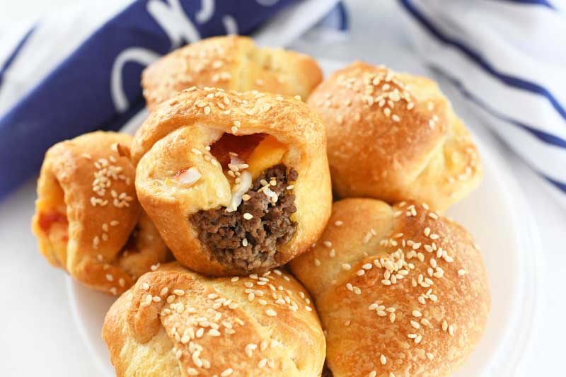 Cheeseburger Crescent Dough Bites are on a white plate with a blue and white napkin in the background. Hamburger is peeking out of the cups. 