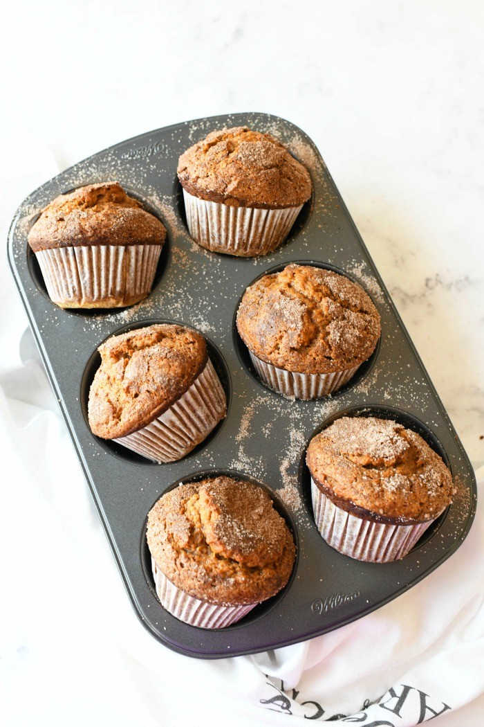 Jumbo Pumpkin Muffins with Cinnamon + Sugar in a grey muffin tin. There is a white baking towel nearby.