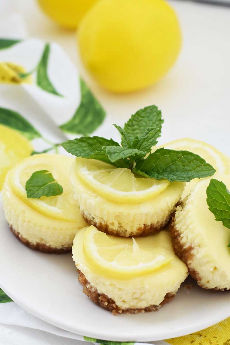 Mini Lemon cheesecakes are stacked on a white plate. These muffin tin cheesecakes have mint and lemon slices on them. 