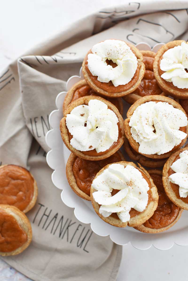 Muffin Tin pumpkin pies are served with whipped cream and cinnamon on a white, scalloped cake stand. 