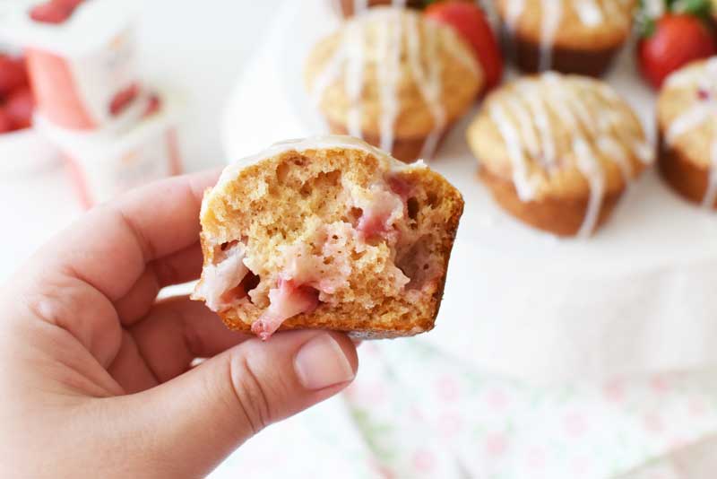 A halved strawberry yogurt muffin is in a white hand. There are muffins in the background on a white cake stand.