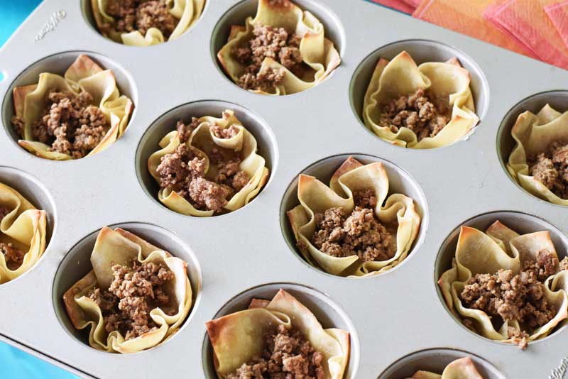 Taco meat is filled in baked wonton cups. They are in a silver muffin tin on a blue table.