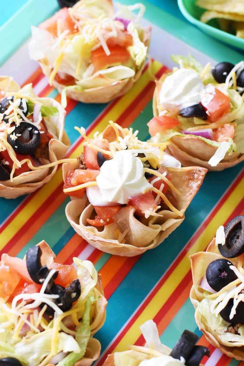 Wonton Taco cups are loaded with filling like ground beef, sour cream, cheese, olives, lettuce, and more! They are on a multi-colored, striped platter on a blue table.