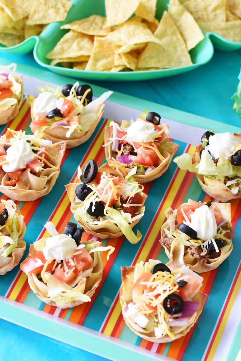 Fully loaded wonton tacos are on a striped, multi-colored platter. There are blue bowls with corn chips in the background.