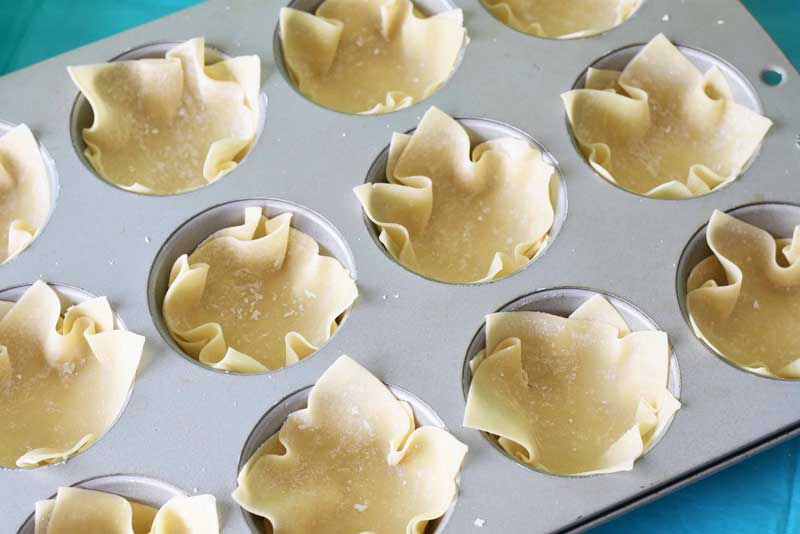 Baking double layers wonton wrappers in a muffin tin. The tin is silver and the wrappers are golden colored on a blue table.