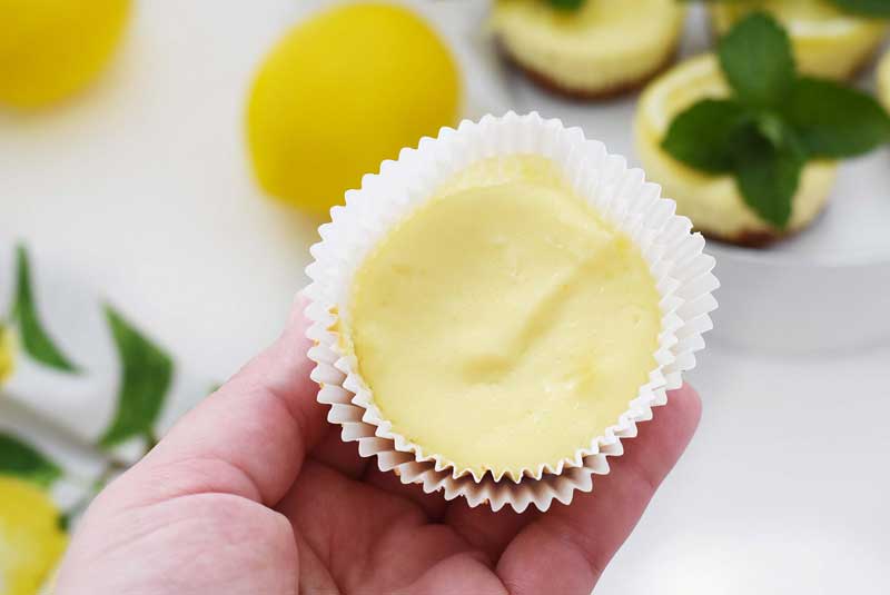 Mini lemon cheesecake in a white double liner in someone's hand. There are lemons and mini cheesecakes in the background. 