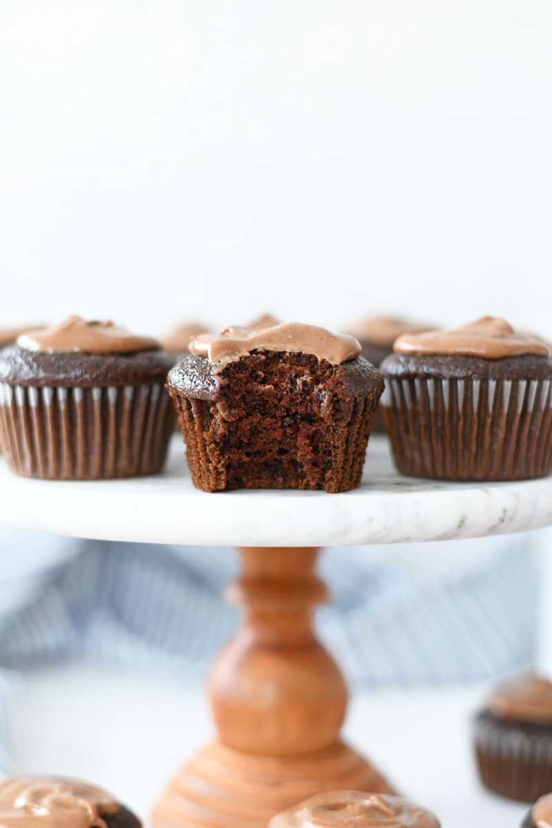 Chocolate frosted cupcakes are on a marble and wood cake stand. A bite is taken out of the middle of the cake.