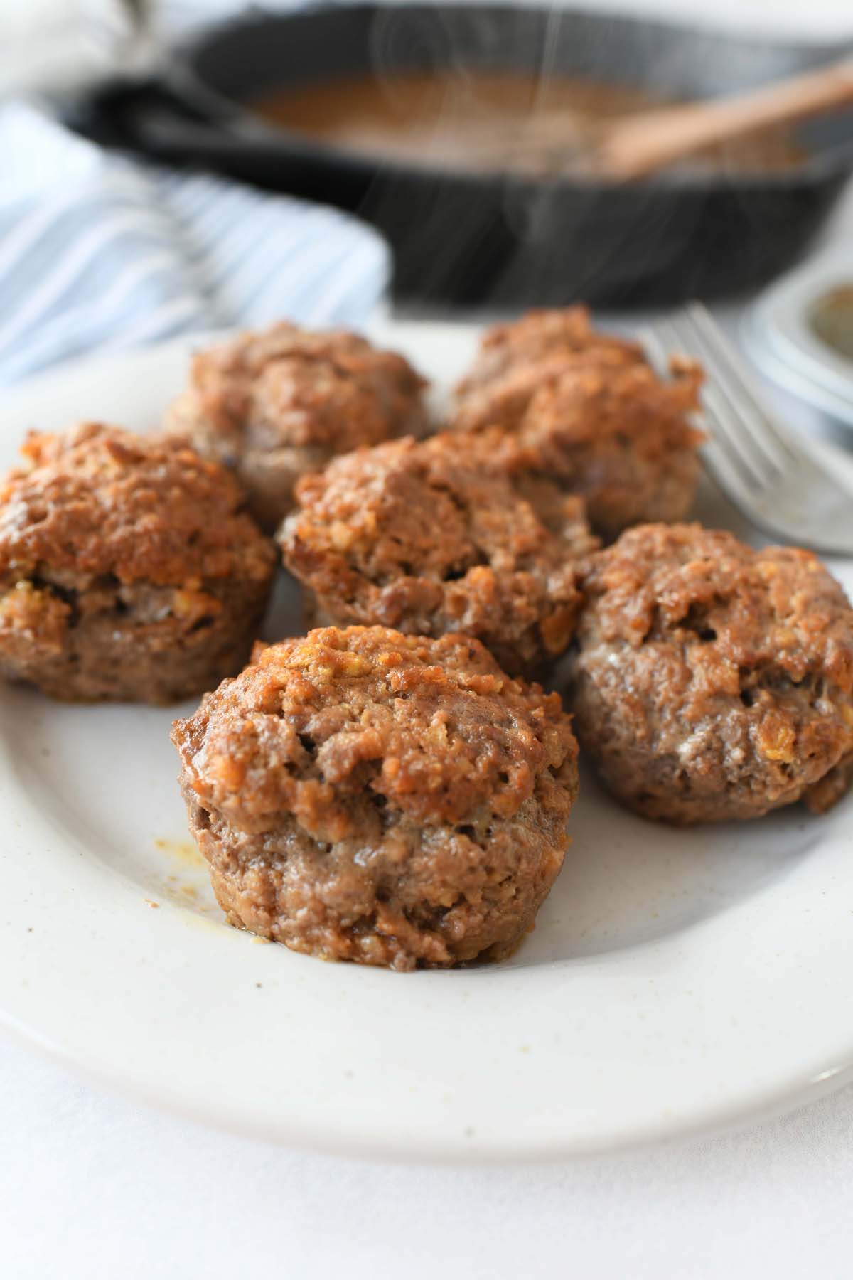 Muffin Tin Steak Patties on a white plate with a simmering pan of gravy in the background. 