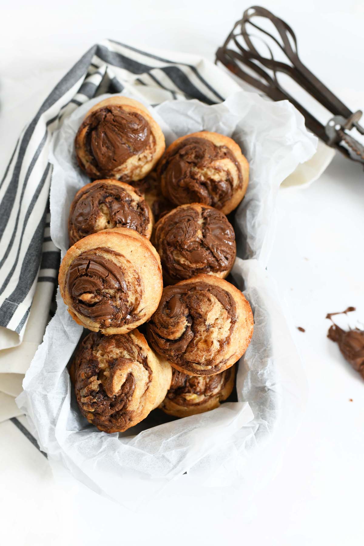 Nutella Banana Swirl Muffins in a parchment-lined pan. There is an old-fashioned hand mixer in the background and a grey and tan striped napkin.