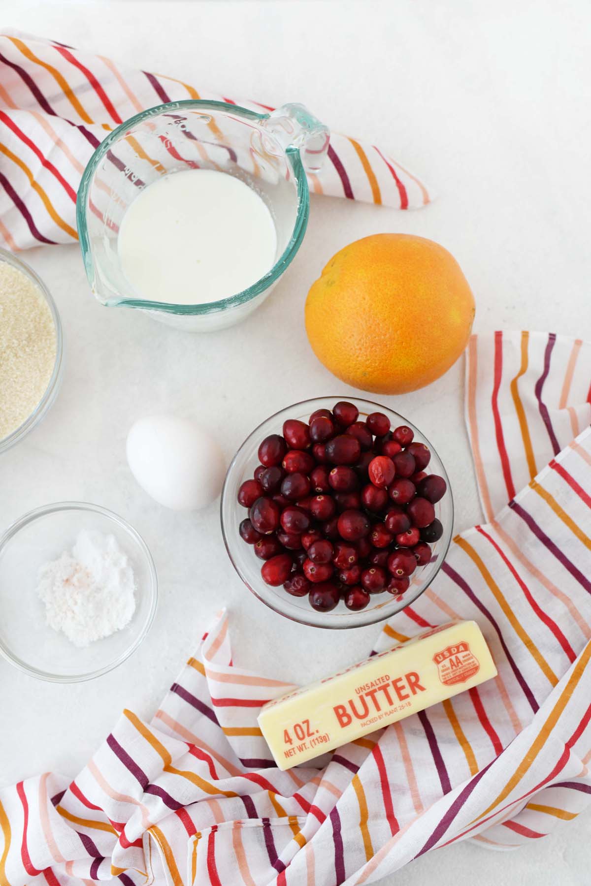 Orange cranberry muffin ingredients on a white table with a colorful striped napkin.