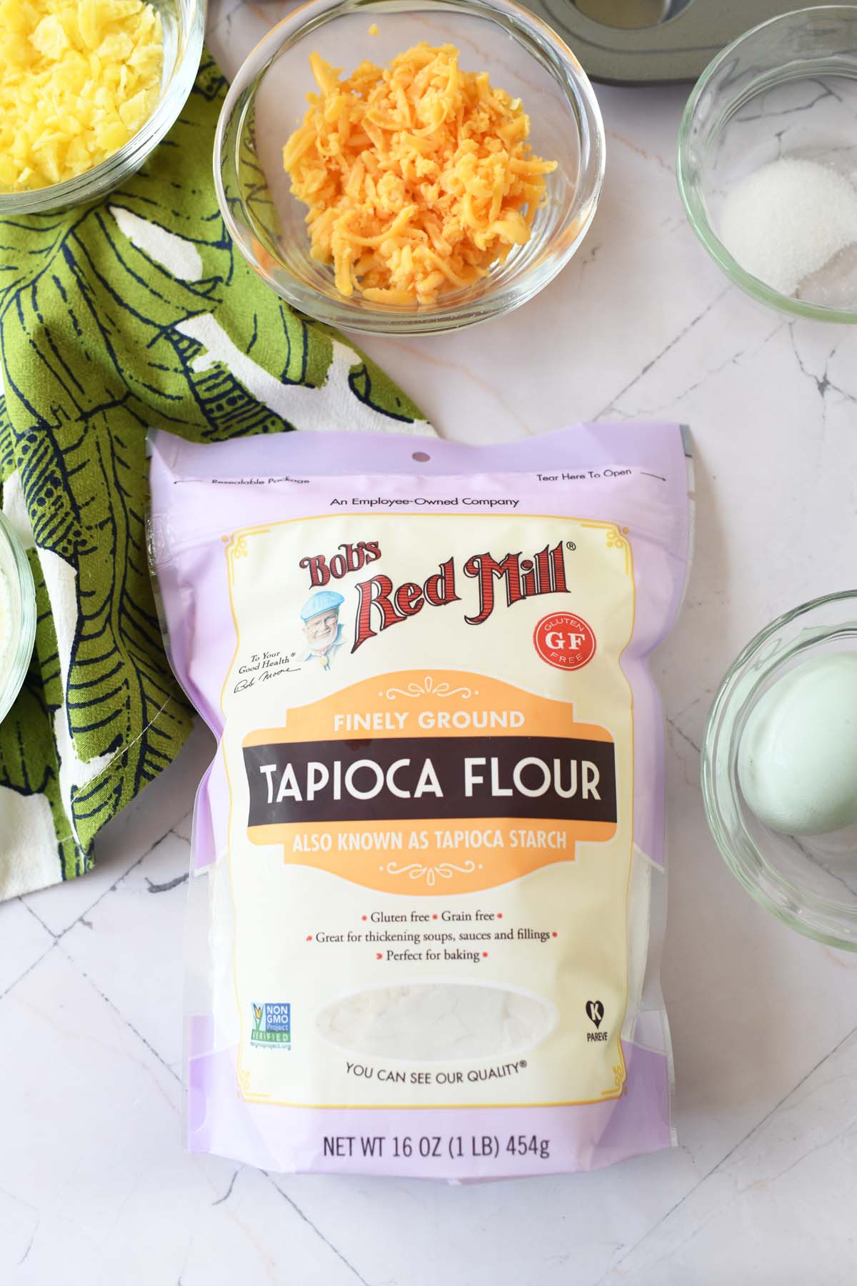 A bag of Bob's Red Mill Tapioca Flour on a white surface near other ingredients in glass bowls.