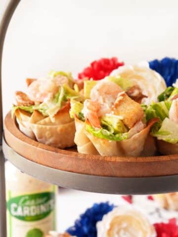 An upclose shot of shrimp caesar salad wonton cups on a wooden tray near red, white, and blue flowers.