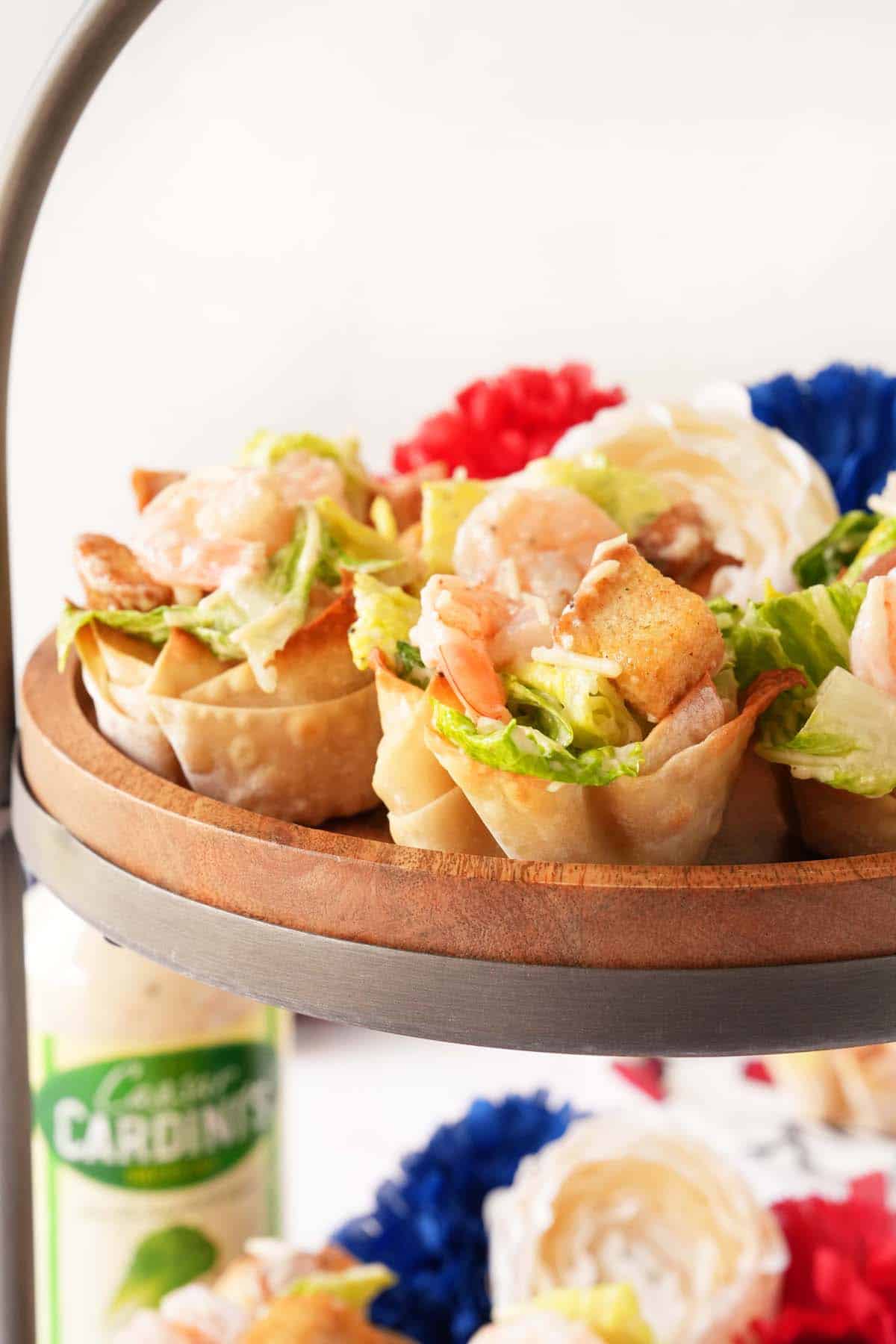 An upclose shot of shrimp caesar salad wonton cups on a wooden tray near red, white, and blue flowers.