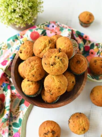 Golden-brown mini chocolate chip muffins in a wooden bowl near a floral napkin with a green plant nearby.