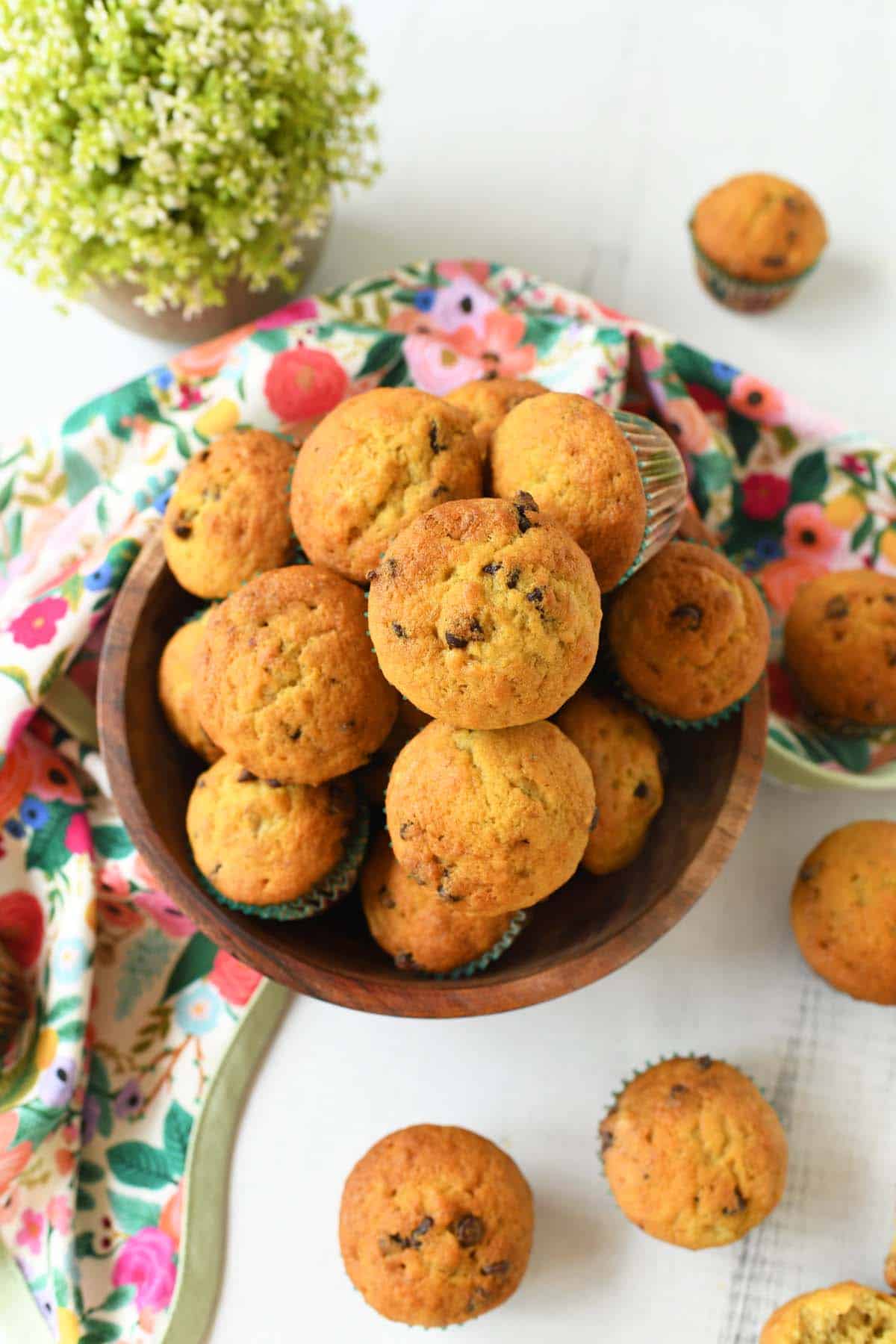 Golden-brown mini chocolate chip muffins in a wooden bowl near a floral napkin with a green plant nearby.