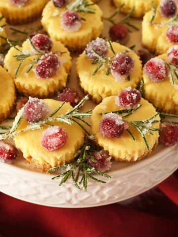Mini festive cheesecakes with sugared cranberries and rosemary on a platter.