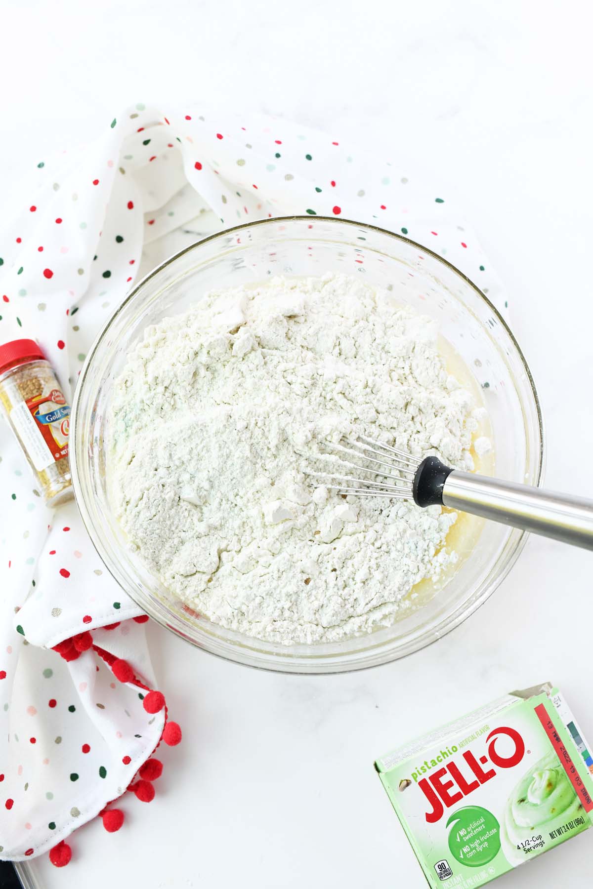 A bowl with a silver whisk, and dry ingredients. A dotted napkina nd box of Jello-0 pistachio pudding are nearby.