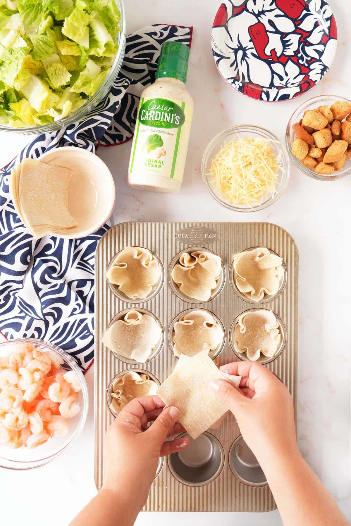 Hands forming wonton cups in a silver muffin tin near ingredients.