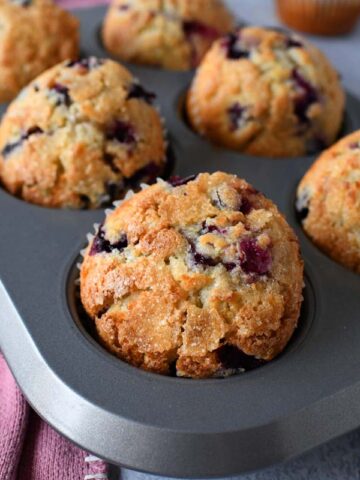 An overhead view of a metal muffin pan holding six domed, sugar-dusted blueberry muffins in parchment liners. A deep purple cloth napkin is placed beside the tin on a grey surface.