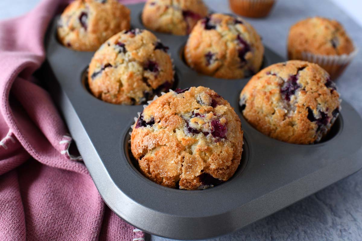 An overhead view of a metal muffin pan holding six domed, sugar-dusted blueberry muffins in parchment liners. A deep purple cloth napkin is placed beside the tin on a grey surface.